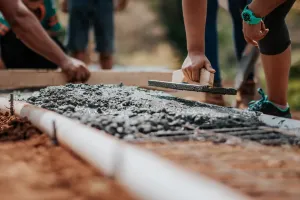 Model drewnianego domku na betonowym fundamencie leży na planach budowy obok miarki i ołówka, symbolizując koszt fundamentu.